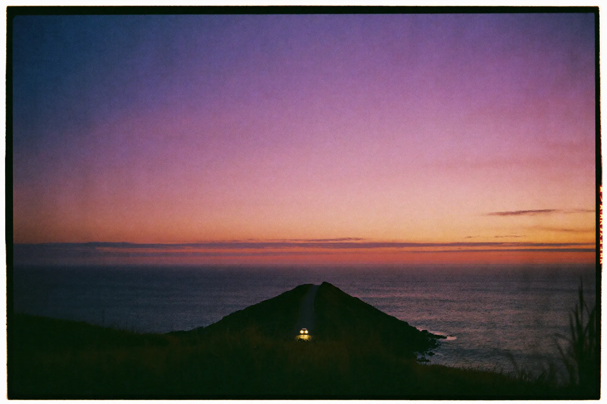 Coastal road hugging low cliffs above a calm sea at dusk, one car with headlights on, violet and magenta sky