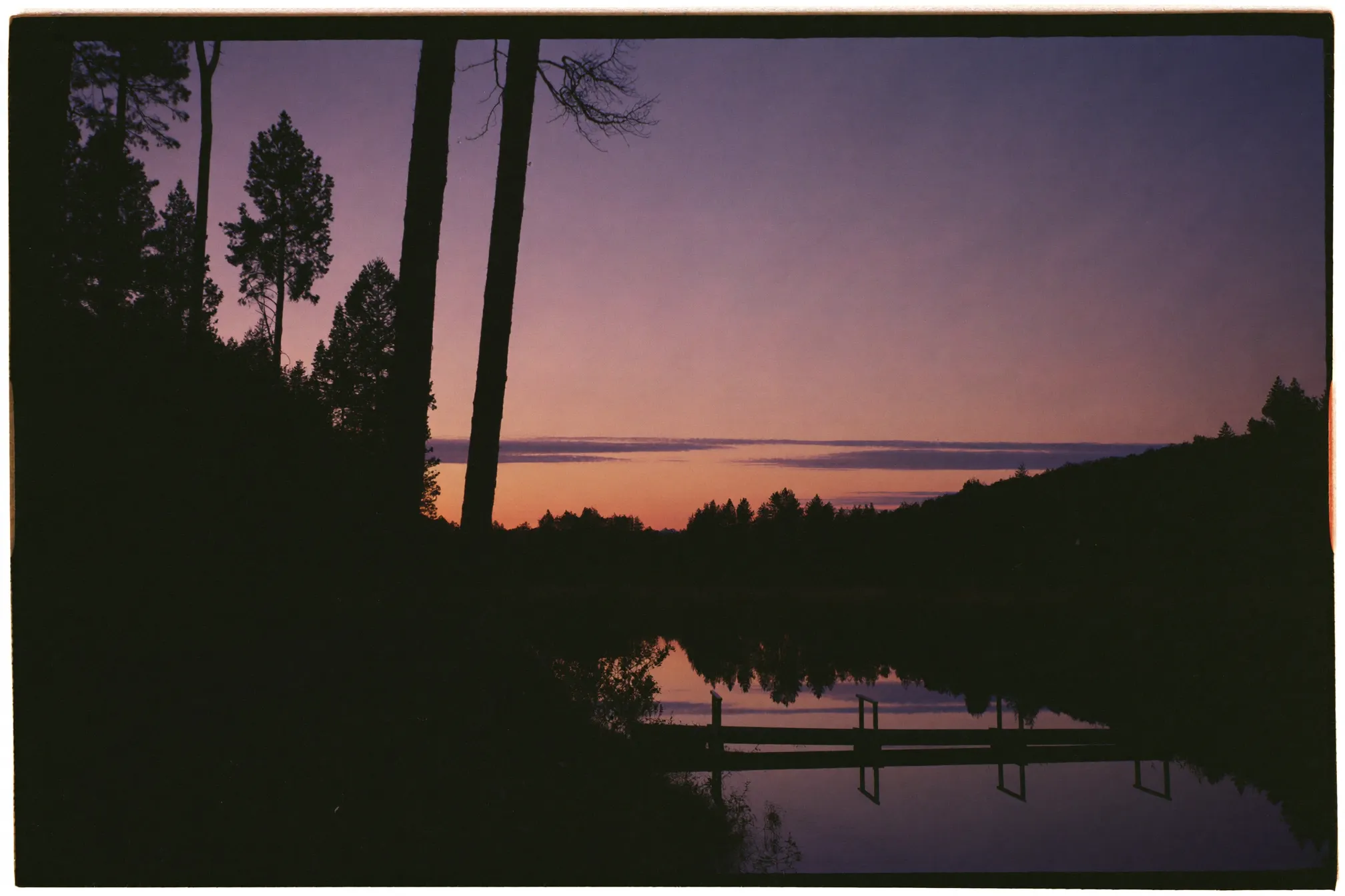 Quiet forest road beside a still lake at dusk, pine silhouettes and a wooden jetty reflected in violet water